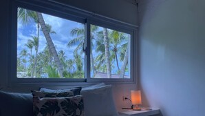 A bedroom with a bed, pillows, a nightstand, a lamp, and a window with a view of palm trees. - Hotel Boutique Casa Grande Playa Bonita (Las Terrenas)