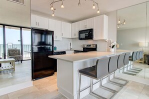 A modern kitchen with white cabinets, a black refrigerator, a microwave, and a bar area with chairs. - Oceanfront 2-Bedroom Condo in Carolina Beach 0512 (Carolina Beach)