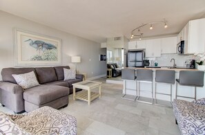 Una sala moderna con un sofá, una mesa de centro y un pouf estampado. Se ve una cocina con gabinetes blancos y un refrigerador al fondo.