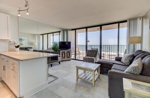 A modern living room with a sofa, a coffee table, a TV, and a balcony with a view. - Oceanfront 2-Bedroom Condo in Carolina Beach 0512 (Carolina Beach)