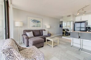 A living room with a sofa, a coffee table, and a kitchen area with a refrigerator and a dining table. - Oceanfront 2-Bedroom Condo in Carolina Beach 0512 (Carolina Beach)