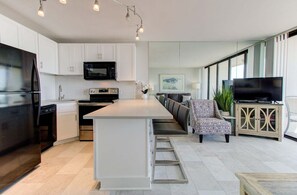 Una cocina moderna con armarios blancos, un refrigerador negro, un microondas y una estufa. Hay un comedor con una mesa y sillas blancas, y un televisor sobre un mueble de madera.