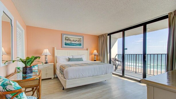 Una camera da letto con un letto, una sedia, un comodino, una lampada, una pianta e un balcone con vista sull'oceano.