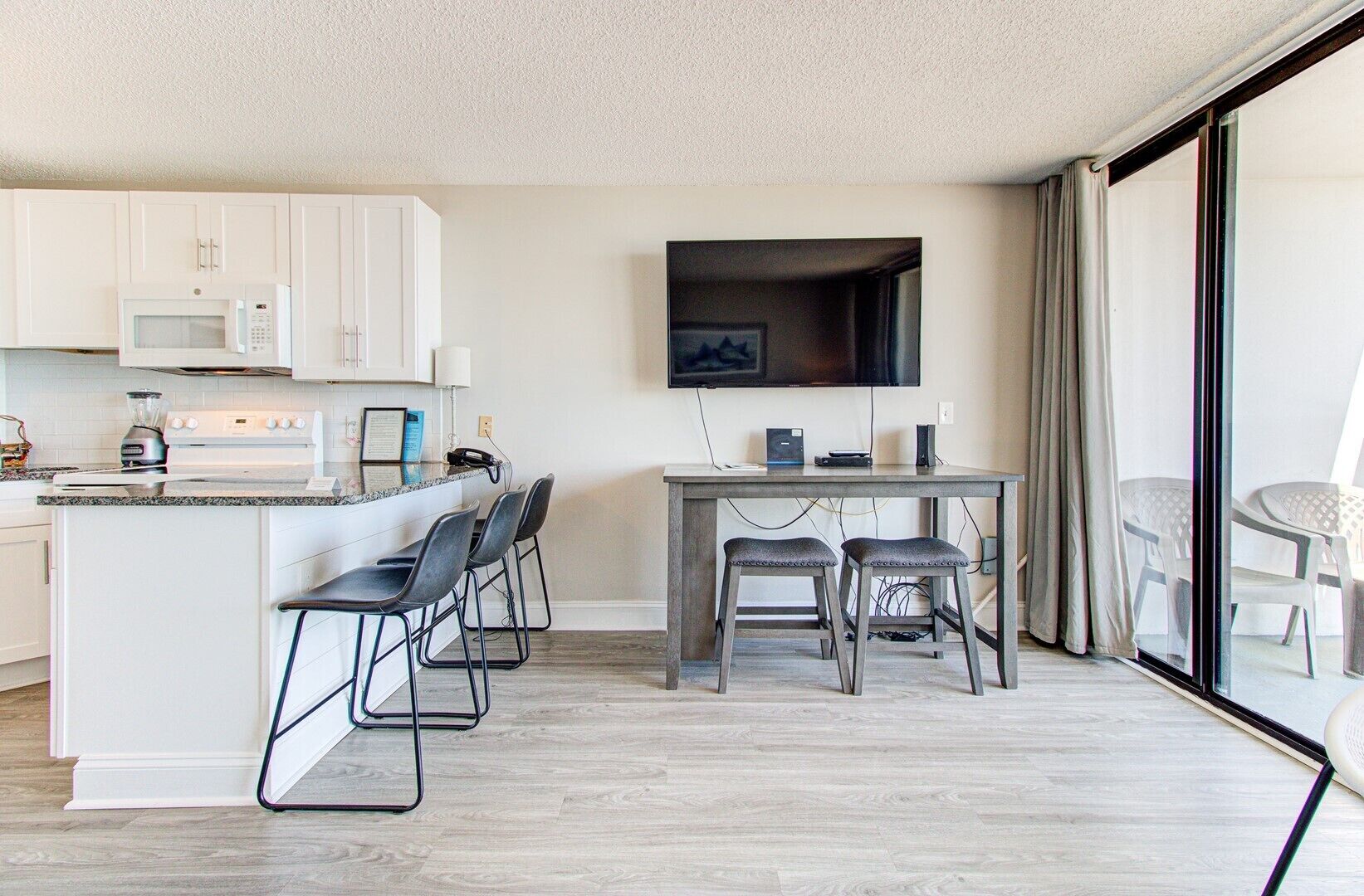 A modern kitchen with white cabinets, a microwave, and a stove. There is a dining area with a table and chairs, and a TV mounted on the wall.