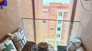 Un balcón con barandilla de vidrio, un banco tejido, una mesa pequeña con un cuenco y vista a un edificio de varios pisos.