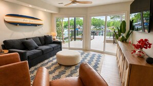 A modern living room with a sofa, ottoman, and leather chairs, a ceiling fan, and a view through large sliding glass doors to an outdoor deck. - Makua Jim’s - Quiet Cove Retreat with Swim Dock, 3 Decks, boat slip, & game room (Sunrise Beach)