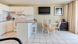 A compact kitchen with white cabinets, a microwave, and a small island with stools. A flat-screen TV is mounted on the wall, and there is a framed picture above it. - Breathtaking oceanfront views await at 0610 Smooth Sailing in Carolina Beach. (Carolina Beach)
