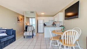 A compact living space with a kitchenette, a dining area with white chairs, a sofa, and a flat-screen TV mounted on the wall. - Breathtaking oceanfront views await at 0610 Smooth Sailing in Carolina Beach. (Carolina Beach)