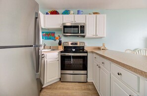 A compact kitchen with white cabinets, a stainless steel oven, a microwave, and a refrigerator. - Enjoy oceanfront bliss in 0612 Sandy Feet Retreat, a 2BR condo in Carolina Beach. (Carolina Beach)