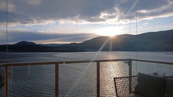 A view from a boat deck with a railing, a body of water, and distant mountains under a cloudy sky with the sun setting or rising.