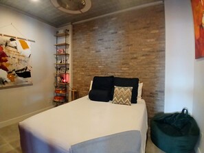 Un dormitorio con cama, pared de ladrillo, estante con objetos y una obra de arte colgada.