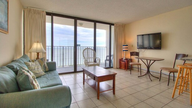 Una habitación con sofá, mesa, sillas, televisión y un balcón con vistas.