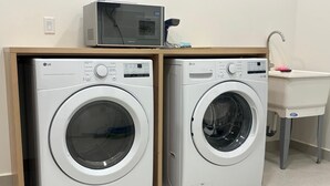 Deux lave-linge à chargement frontal blancs de la marque LG se trouvent dans un espace buanderie. Un micro-ondes est posé sur le dessus d’une armoire en bois, et un évier avec un robinet est également présent.