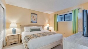 A hotel room with a bed, two bedside tables with lamps, a window with curtains, and a television on a stand. - Oceanfront charm awaits at 0709 Seaside Serenade in Carolina Beach! (Carolina Beach)