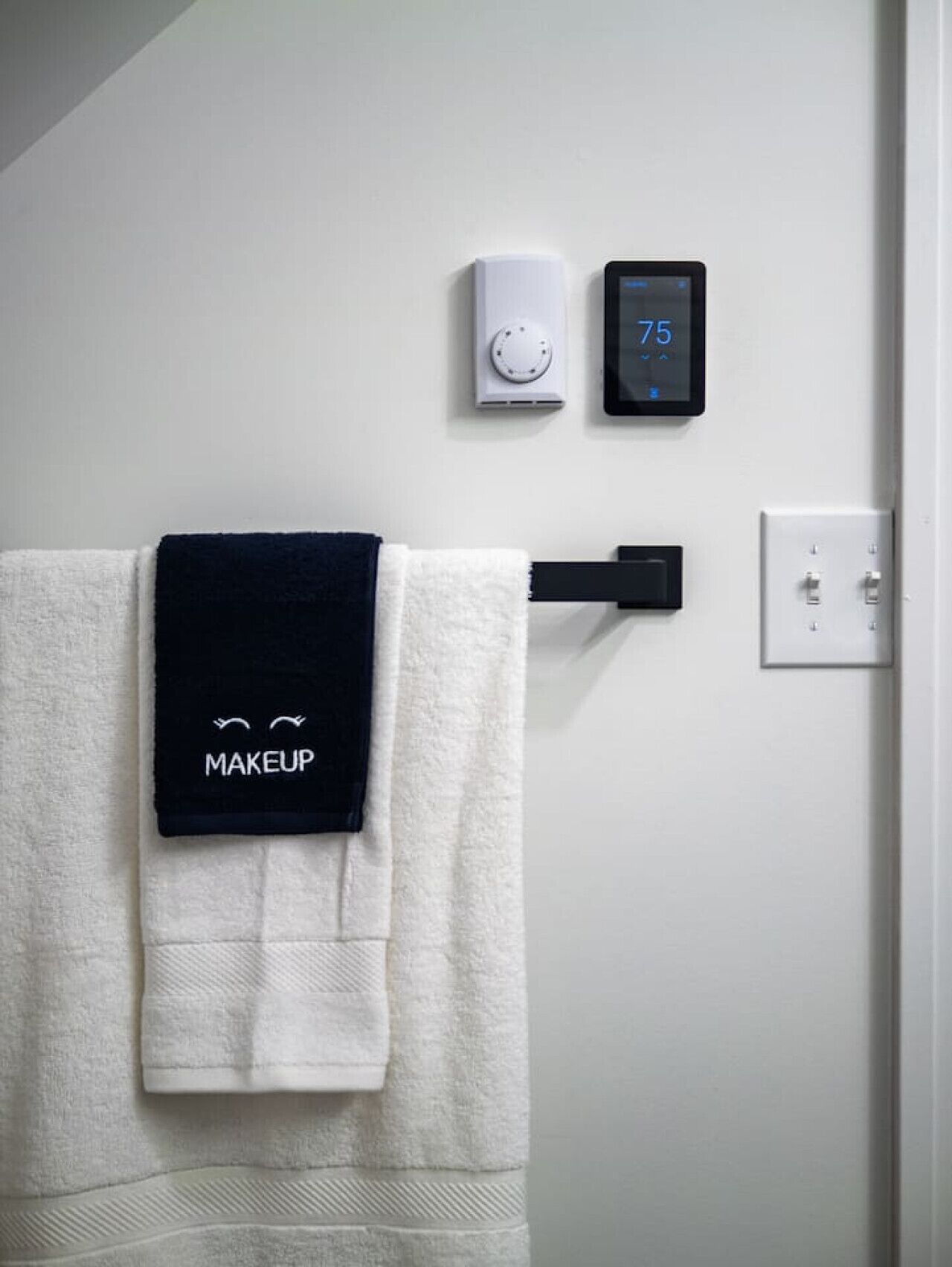 Un baño con un perchero que sostiene una toalla blanca y una toalla negra con el texto 'MAKEUP'. Se ve un termostato digital y un dispositivo montado en la pared.