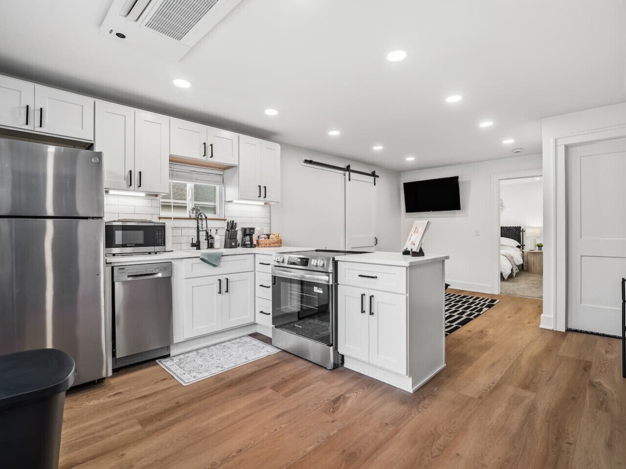 A modern kitchen with white cabinets, stainless steel appliances, a flat-screen TV, and wooden flooring.