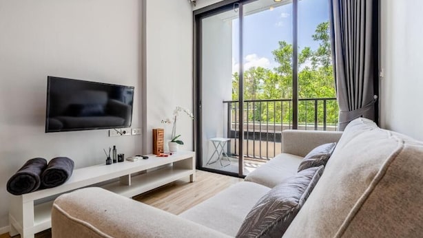 A modern living room with a flat-screen TV, a sofa, a small white table, and a balcony with a view of trees. - ZcapeX2 (Choeng Thale)