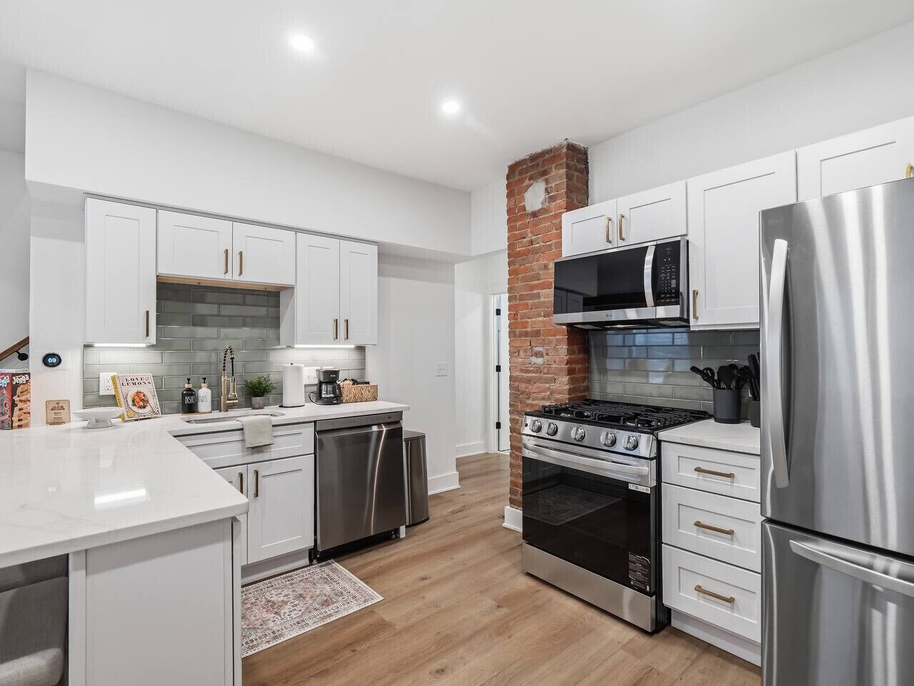 A modern kitchen with white cabinets, stainless steel appliances, a brick accent wall, and a wooden floor.