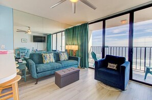 A living room with a sofa, ottoman, and a chair, a television, a ceiling fan, and a view of the ocean through large windows. - Oceanfront 2BR condo for 6 in Carolina Beach 0712 (Carolina Beach)