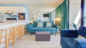 Ett modernt kök med vita skåp, en matplats med en blå soffa och trästolar samt ett stort fönster med gröna gardiner.