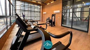 A gym with exercise equipment, large windows, and a brick wall. - Vol 29 - Miramar Hospedagens (João Pessoa)