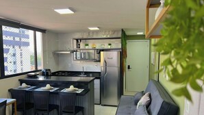 A compact kitchen with a central island, a microwave, a stainless steel refrigerator, and a dining area with chairs and a table. - Vol 29 - Miramar Hospedagens (João Pessoa)