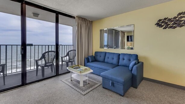 A balcony with a view of the ocean, a blue sofa, a small table with a tray, and a mirror on the wall. - Oceanfront 1-bedroom condo in Carolina Beach 0806 (Carolina Beach)