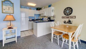 A compact kitchen with white cabinets, a central island, a dining table with chairs, a lamp, and wall decorations including a clock and a motivational sign. - Oceanfront 1-bedroom condo in Carolina Beach 0806 (Carolina Beach)