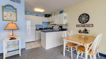 Een compacte keuken met witte kasten, een centraal eiland, een eettafel met stoelen, een lamp en wanddecoraties, waaronder een klok en een motiverende tekst.