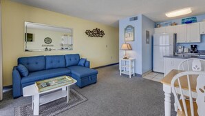 A living room with a blue sofa, a coffee table, a lamp, and a kitchen area with white cabinets and appliances. - Oceanfront 1-bedroom condo in Carolina Beach 0806 (Carolina Beach)