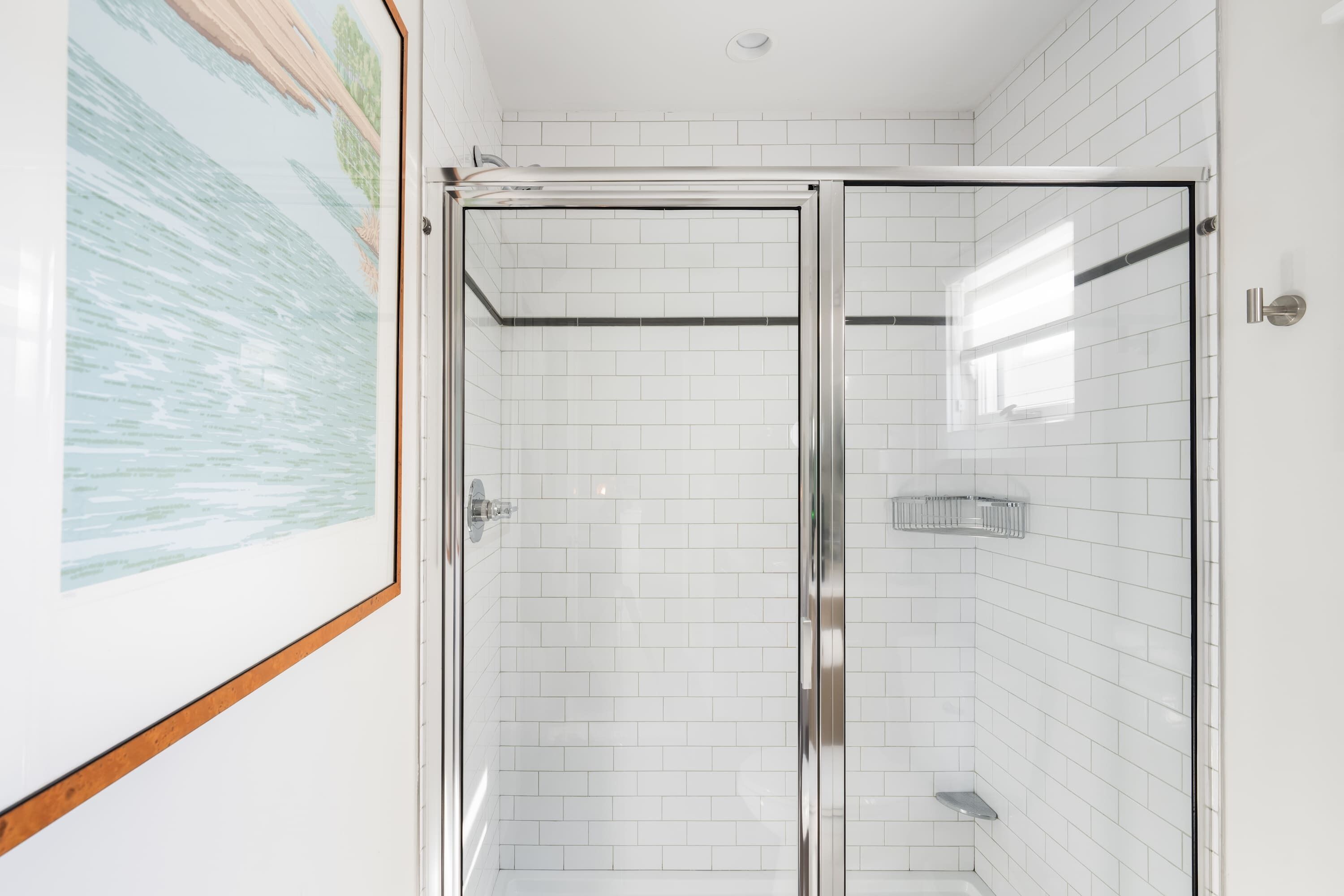 Un baño moderno con paredes de azulejos blancos, cabina de ducha de vidrio e imagen enmarcadas en la pared.