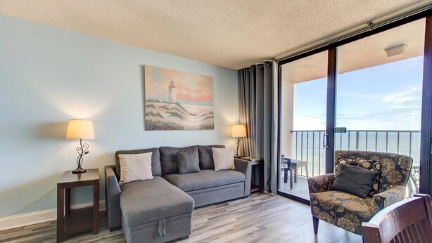 A room with a sofa, a chair, a painting, and a view of the sea. - Cozy 1 Bedroom Oceanfront Condo at Carolina Beach 0809 (Carolina Beach)