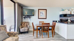 A room with a dining table, chairs, a sofa, a TV, and a kitchen area with appliances. - Cozy 1 Bedroom Oceanfront Condo at Carolina Beach 0809 (Carolina Beach)