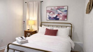 A hotel room with a bed, a nightstand with a lamp, a painting on the wall, and a window with curtains. - Spacious 4BR Family Home with Game Room (Raleigh)