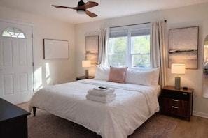 A hotel room with a bed, two bedside tables, a ceiling fan, a window with curtains, and artwork on the walls. - Spacious 4BR Family Home with Game Room (Raleigh)