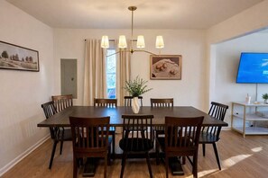 A dining room with a large table, six chairs, a chandelier, a vase with plants, a painting, and a television on the wall. - Spacious 4BR Family Home with Game Room (Raleigh)