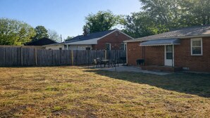 A backyard with a wooden fence, a patio with chairs, and a small brick building. - Spacious 4BR Family Home with Game Room (Raleigh)