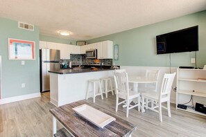 A compact kitchen with white cabinets, a microwave, and a sink. There is a dining area with white chairs and a table. A flat-screen TV is mounted on the wall. - Oceanfront 2-Bedroom Escape in Carolina Beach - Perfect for 6! (Carolina Beach)