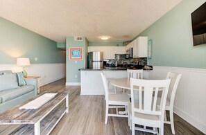 A compact living space with a kitchenette, a dining area with white chairs, a sofa, and a coffee table. - Oceanfront 2-Bedroom Escape in Carolina Beach - Perfect for 6! (Carolina Beach)