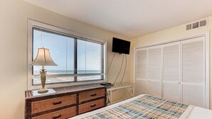 Ein Schlafzimmer mit einem Bett, einer Kommode mit Schubladen, einer Lampe, einem an der Wand montierten Fernseher und einem Fenster mit Jalousien.
