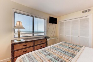 A bedroom with a bed, a dresser with drawers, a lamp, a TV mounted on the wall, and a window with blinds. - Experience oceanfront bliss at 0901 Salty Dayz Tower 0901 (Carolina Beach)