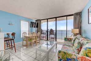 Ein Zimmer mit Balkon, einem Essbereich mit Stühlen und Tisch, einer Couch mit farbenfroher Auflage und einem an der Wand befestigten Fernseher.