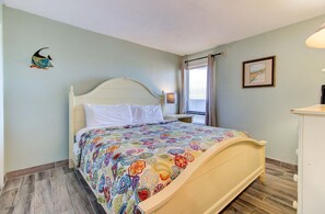 A bedroom with a bed, a nightstand, a lamp, a window with curtains, and a wall-mounted picture. - Experience oceanfront bliss at 1002 in Carolina Beach, perfect for 4 guests. (Carolina Beach)
