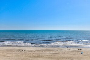 Una playa con cielo despejado, mar tranquilo y algunas personas caminando.