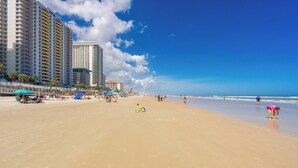 Ein weiter Sandstrand mit Hochhäusern im Hintergrund, Sonnenschirmen und Menschen.
