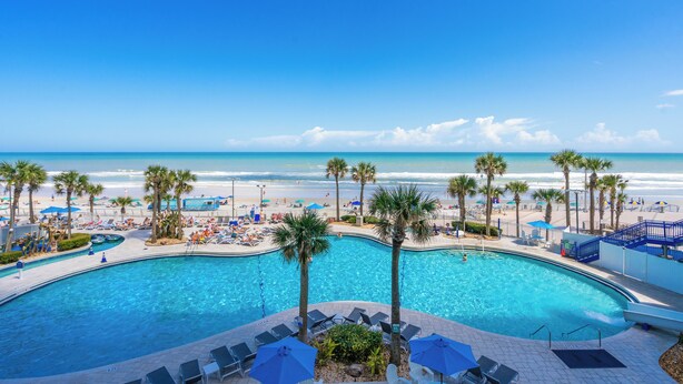 Ein Swimmingpool mit Liegestühlen und Sonnenschirmen, ein Strand und Palmen.