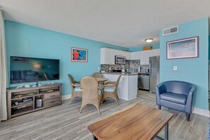 A living room with a TV, a dining area with wicker chairs, a kitchen with white cabinets, and a blue chair. - Oceanfront Premier 1 Bedroom Condo at Carolina Beach, perfect for a serene getaway! (Carolina Beach)