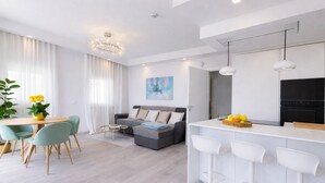 Ein modernes Wohnzimmer mit Sofa, einem Esstisch mit Stühlen, einer Küchenzeile mit Bar und einem großen Fenster mit Vorhängen.