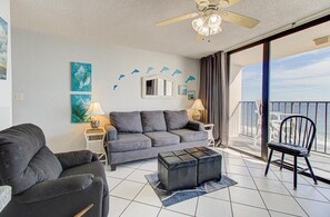 Een woonkamer met een bank, een fauteuil, een salontafel, een plafondventilator en een balkon met uitzicht.