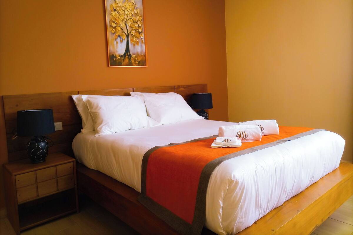A hotel room with a large bed, two bedside tables, a painting on the wall, and a red accent on the bedspread.